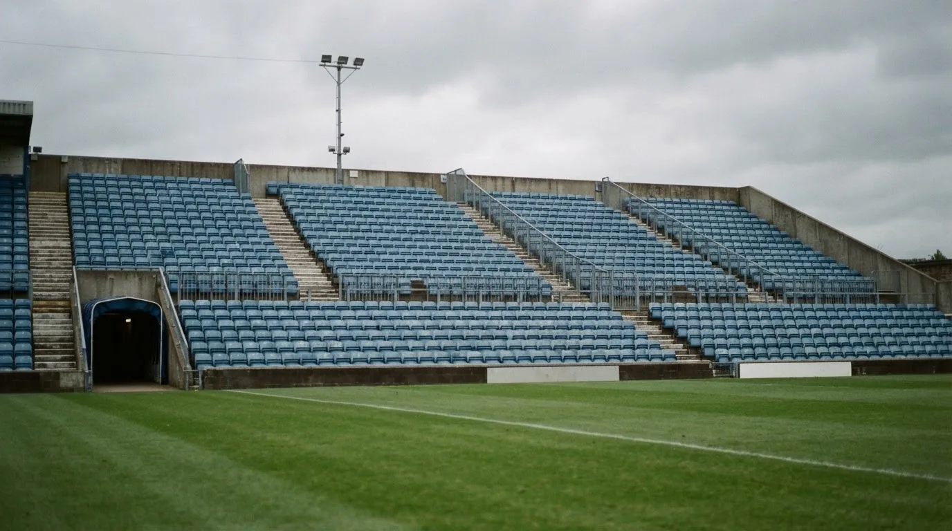Rzędy pustych plastikowych krzesełek małego stadionu widziane z poziomu murawy pod pochmurnym niebem.