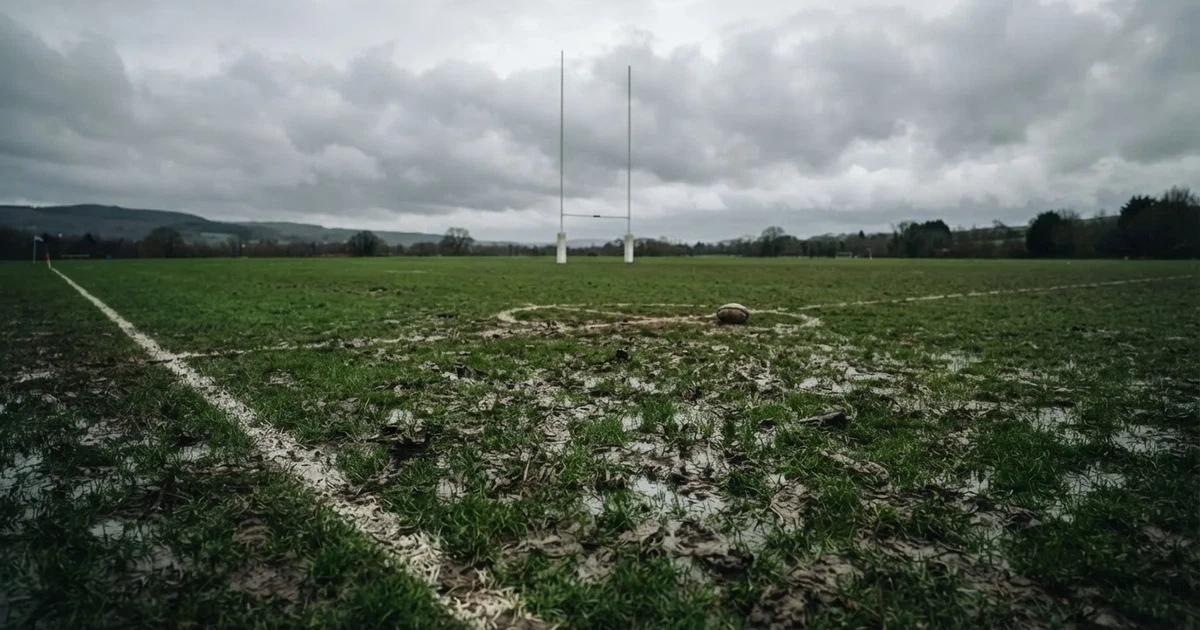 Boisko do rugby ze słupami bramki w oddali pod pochmurnym niebem, mokra trawa i ślady butów.