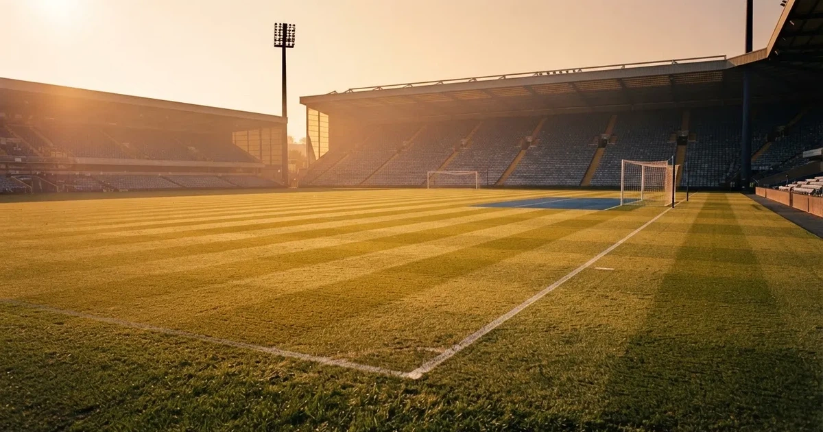Murawa stadionu o zachodzie słońca z długimi cieniami słupków bramki, trybuny w ciepłym świetle.