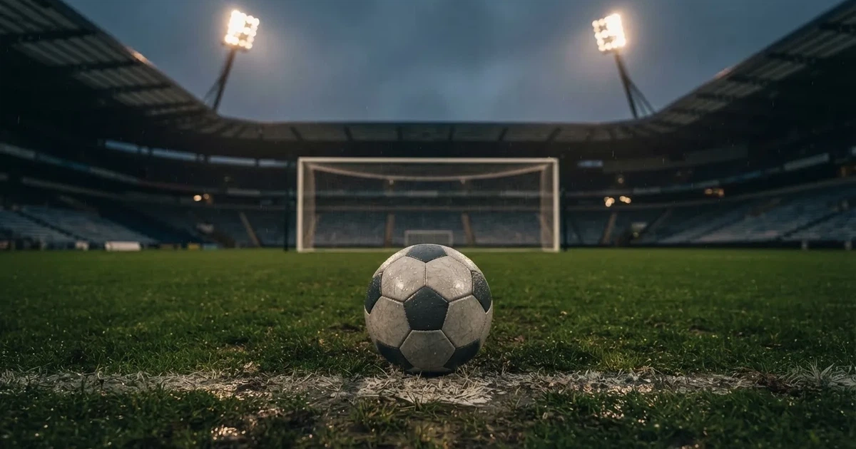 Futbolowa piłka stoi na punkcie karnym w pustym stadionie podczas mżawki, w tle rozmyte trybuny i jasne światło masztów.