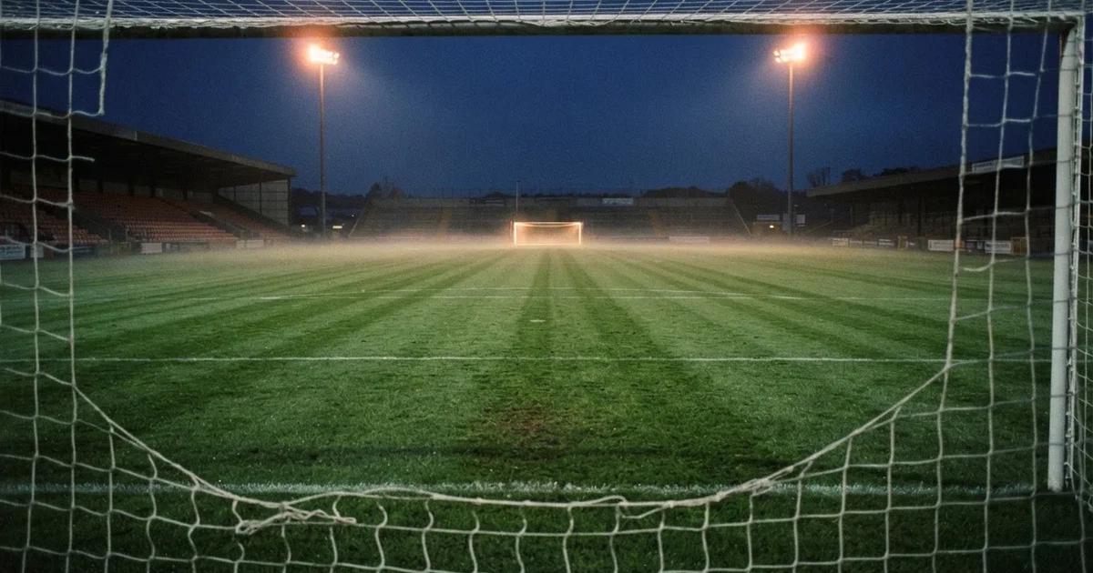 Pusta murawa stadionu miejskiego w chłodny jesienny wieczór, siatka bramki świeci w świetle reflektorów, nad boiskiem wisi mgła.