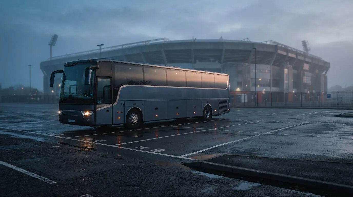 Autokar stoi na parkingu przed stadionem o świcie, rozmyte kontury trybun w tle, pusty plac i mokry asfalt.